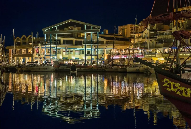 Benalmadena marina nightlife, lit terrace bars on the inner harbour promenade at night with groups of people and coloured reflections in the water
